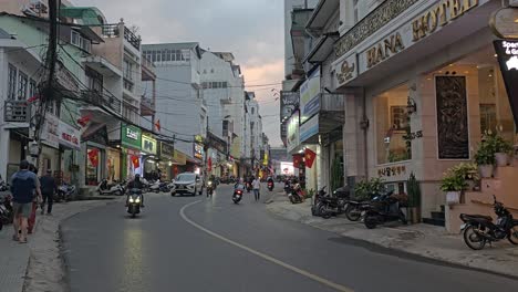 Motos-Y-Automóviles-Circulando-Por-La-Calle-A-Lo-Largo-De-Las-Tiendas-Y-El-Hotel-Al-Atardecer-En-La-Ciudad-De-Da-Lat,-Vietnam