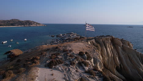 Greek-Country-Flag-Waving-on-Pole-on-Cape-by-Aegean-Sea,-Revealing-Drone-Shot