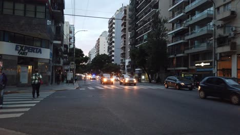 Buses-taxis-and-traffic-driving-at-buenos-aires-city-pedestrians-walk-at-corrientes-avenue,-sunset-skyline-between-metropolitan-buildings-and-stores