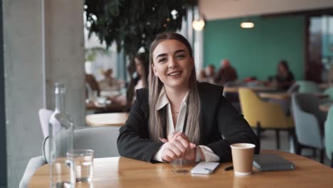 Encantadora-Joven-Sentada-En-Un-Café-Vestida-De-Negocios-Con-Una-Sonrisa,-Mirando-A-La-Cámara