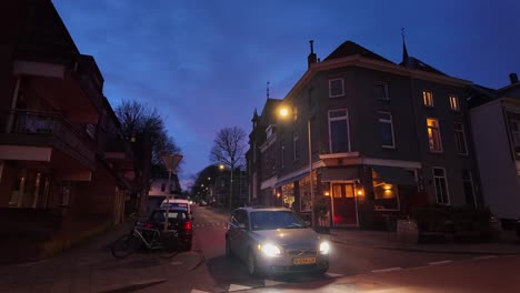Car-with-lights-on-driving-through-Dutch-street-at-night-in-Arnhem-wide-shot