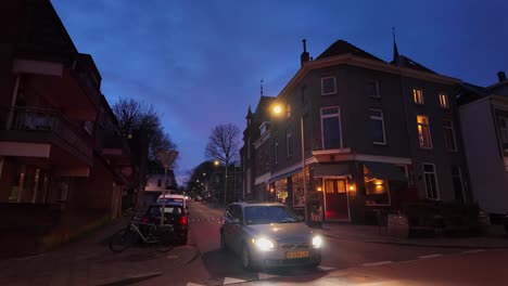 Coche-Con-Luces-Encendidas-Conduciendo-Por-La-Calle-Holandesa-Por-La-Noche-En-Arnhem,-Plano-Amplio.