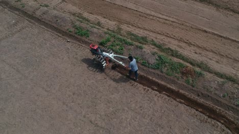 Disparo-Desde-Un-Dron-Volando-Sobre-Un-Tractor-Con-Sistema-De-Grada-Arando-El-Suelo-En-Un-Campo-Agrícola-Cultivado,-Pilar-De-Senderos-De-Polvo-Detrás,-Preparando-El-Suelo-Para-Plantar-Nuevos-Cultivos,-Concepto-De-Agricultura,-Vista-Superior