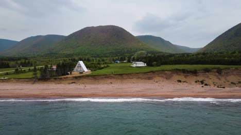 Aerial-drone-view-revealing-the-incredible-scenery-of-Cape-Breton-Island-in-Nova-Scotia-where-an-A-Frame-cabin-right-on-a-cliffs-edge-with-waves-crashing-on-the-beach-and-mountains-in-the-background