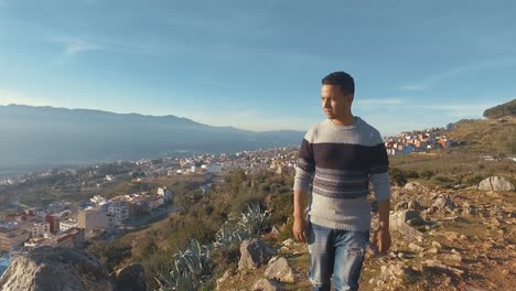 Camera-follows-young-Moroccan-man-walking-up-on-top-of-mountain-summit,-Chefchaouen-Morocco,-sunny-day