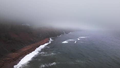 Aerial-drone-shot-panning-upwards-a-beautiful-scene-off-the-coast-of-Cape-Breton-in-Nova-Scotia-where-the-Atlantic-Ocean-is-crashing-onto-giant-cliffs-with-a-waterfall-gushing-into-the-ocean