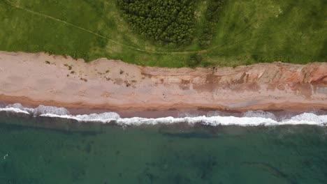 Drone-shot-going-to-the-left-following-the-beautiful-contrast-of-where-the-massive-cliffs-meet-the-beach-and-the-Atlantic-Ocean-located-on-Cape-Breton-Island-in-the-province-of-Nova-Scotia,-Canada