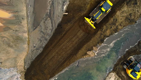 Aerial-top-down-zoom-out-shot-Bulldozer-carrying-dirt-and-excavator-on-the-bank-of-a-stream-removing-sediments