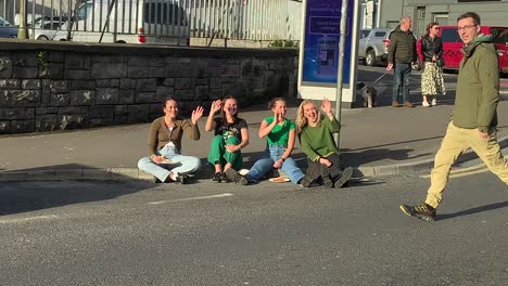 Cheerful-girls-sitting-on-a-sidewalk-wave-to-the-camera-during-Saint-Patrick's-Day-Galway,-Ireland