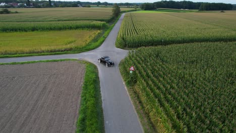 A-black-vintage-car-turns-around-at-an-intersection-next-to-May-fields