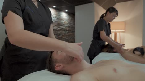 A-couple-gets-a-massage-in-a-spa,-it's-a-duo-massage,-two-professionals-massage-their-heads