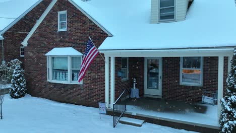 Verschneite-Szene-An-Einem-Backsteinhaus-Mit-Einer-Amerikanischen-Flagge-Und-Einer-Veranda