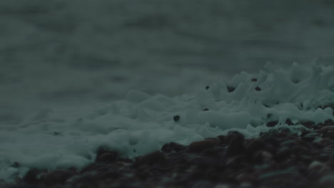Macro-view-of-white-wash-from-foamy-ocean-wave-crashing-and-dispersing-into-dark-grey-pebble-beach