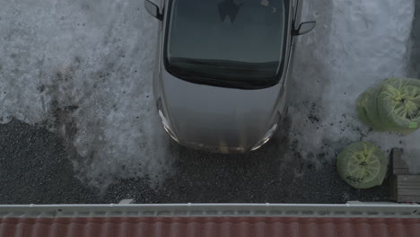 Grey-car-reverses-out-of-garage-onto-ice,-top-down-aerial-view