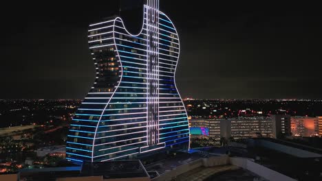 Eine-Luftaufnahme-Des-Gitarrenförmigen-Gebäudes-Des-Seminole-Hard-Rock-Hotel-Und-Casinos,-Das-Nachts-Mit-Neonlichtern-Beleuchtet-Wird-Und-Den-Griechischen-Unabhängigkeitstag-Feiert