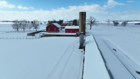 Aerial-view-of-a-rural-snow-covered-landscape-with-a-red-barn,-silo,-and-fence-lines