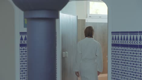 A-woman-in-a-white-bathrobe-in-a-thalassotherapy-center-is-greeted-by-her-masseuse-who-opens-the-door-for-her-massage