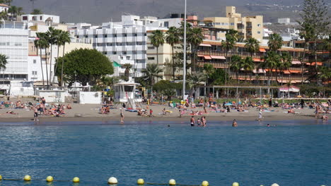 Los-Christianos-Beach---Lively-beach-scene-with-sunbathers-and-swimmers,-framed-by-palm-trees-and-a-backdrop-of-city-buildings