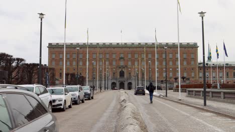 Traffic-in-front-of-Swedish-Royal-Palace-in-Stockholm-on-overcast-day,-flags-flutter-in-wind,-wide-shot