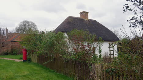 Reetgedecktes-Cottage-Mit-Rustikalem-Zaun-Und-Klassischem-Britischen-Roten-Briefkasten,-Das-Den-Ländlichen-Charme-Englands-In-Einem-Kleinen-Dorf-Auf-Dem-Land-Einfängt