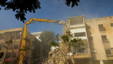 An-Old-Building-Demolition-by-a-Long-Armed-Excavator,-Simultaneously-Employing-Water-Spray-for-Dust-Suppression-in-Tel-Aviv-Jaffa,-Israel---Low-Angle-Shot