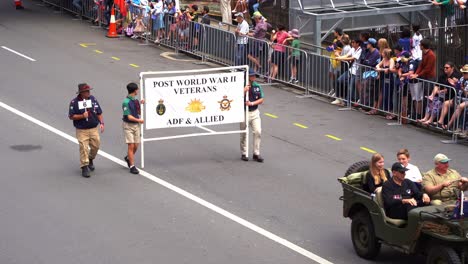 Veteran-and-families-ride-in-a-vintage-military-vehicle-with-a-trailer,-driving-down-the-street,-participating-in-the-Anzac-Day-parade