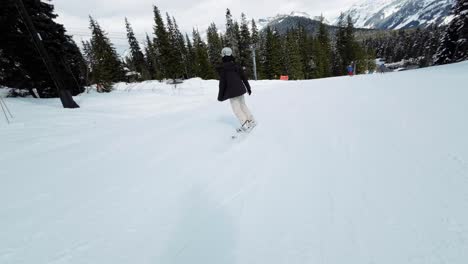 Following-a-tall-Brazilian-adult-woman-dressed-in-black-and-white-snow-gear-snowboarding-in-slow-motion-down-a-ski-resort-path-surrounded-by-pine-trees-at-Snoqualmie-Washington,-USA