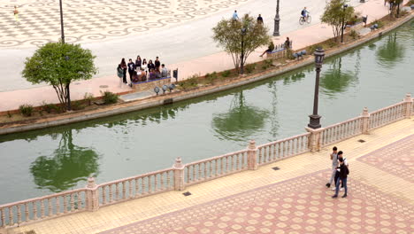 Río-Canal-Con-Gente-Caminando-Sobre-El-Suelo-De-Mosaico-De-Piedra-De-La-Plaza-De-España-En-Sevilla,-Andalucía,-España.