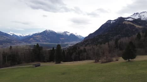 Aerial-view-of-drone-flying-above-huts-on-a-hill-with-snow-capped-mountains-in-background-on-a-cloudy-day-in-Vorarlberg,-Austria