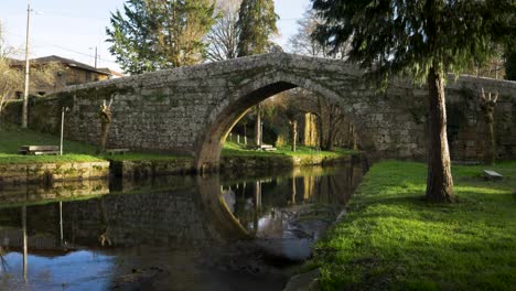 Pan-across-meditative-grassy-river-banks-to-Medieval-roman-bridge-in-Ourense-countryside