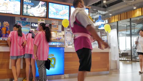 Tres-Jóvenes-Están-Parados-Frente-A-Un-Puesto-De-Comida-Rápida-Mientras-Esperan-Sus-Pedidos-De-Pretzels-Recién-Horneados-En-Un-Centro-Comercial-En-Bangkok,-Tailandia.