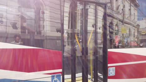 Autobus-drives-streets-of-argentina-south-american-capital-under-sunlight-people-walk-with-wheel-chair,-flores-neighborhood,-pueyrredon-urban-park-greenery-plaza,-Rivadavia-avenue