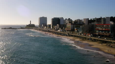 Aerial-view-of-Farol-da-Barra-beach-with-the-people-enjoying-the-sunny-day-and-the-buildings-at-background,-Salvador,-Bahia,-Brazil