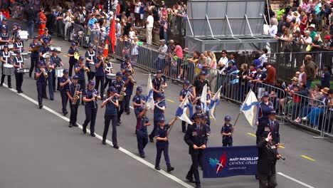 Students-from-the-Mary-MacKillop-Catholic-College-band,-performing-musical-instruments-for-Anzac-Day-parade-with-crowds-lining-the-street-in-downtown-Brisbane-city