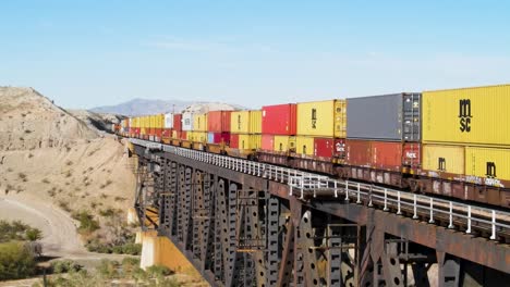 Freight-Train-Carrying-Shipping-Containers,-Multi-Color,-Red,-Yellow,-Over-Steel-Bridge-Between-California-and-Arizona-,-Colorado-River-,-I-40-freeway-East