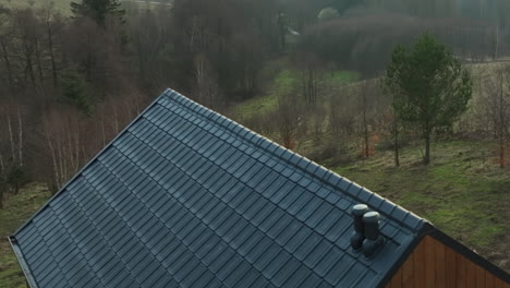 Nahaufnahme-Eines-Schrägen-Dachs-Mit-Grauen-Ziegeln-Auf-Einem-Holzhaus,-Umgeben-Von-Einer-Grasbewachsenen-Landschaft-Mit-Spärlichen-Bäumen
