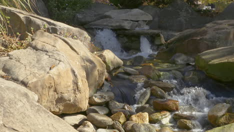 Small-waterfall-flowing-into-fresh-water-stream-and-rocks-of-various-sizes-and-shapes,-during-a-clear-and-sunny-afternoon