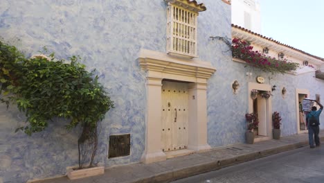 Wall-Shrub-On-Blue-Building-With-Pan-Right-To-Reveal-Old-Town-Street-In-Cartagena-With-People-Walking-Past