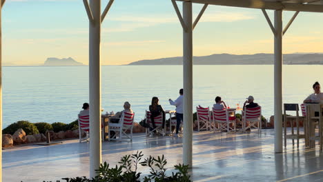 Leute-Sitzen-In-Einem-Restaurant-Mit-Meerblick-Am-Strand-Von-Estepona,-Sonniger-Tag-In-Andalusien,-Spanien,-Sommerurlaubsziel,-Gibraltar-Im-Hintergrund,-4K-Aufnahme