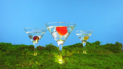 Una-Copa-De-Martini-Apoyada-Sobre-Musgo-Que-Se-Divide-En-Tres-Copas-De-Martini-Con-Varias-Frutas-Que-Se-Dejan-Caer-En-Los-Espíritus-Claros-Del-Interior,-Incluidas-Una-Fresa,-Una-Cereza-Y-Un-Fondo-Azul-Oliva