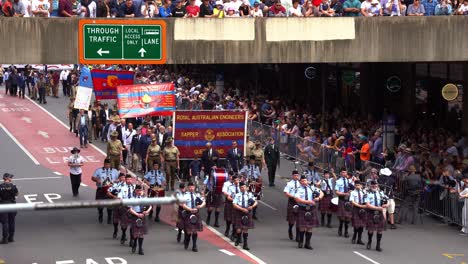 Queensland-Police-Pipes-and-Drums,-pipers-and-drummers,-playing-bagpipes-and-drums-walking-down-the-street,-followed-by-other-units-with-cheering-crowds-at-annual-tradition-of-Anzac-Day-parade