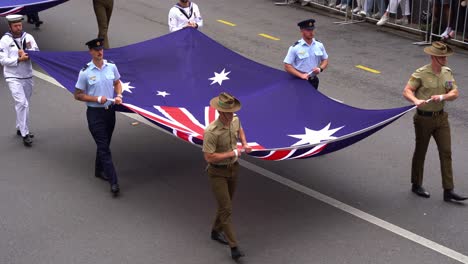 Soldados-De-Las-Tres-Ramas-De-La-Fuerza-De-Defensa-Australiana,-La-Marina-Real-Australiana,-El-Ejército-Australiano-Y-La-Fuerza-Aérea-Real-Australiana-Sosteniendo-Las-Banderas-Marchando-Por-La-Calle-En-El-Desfile-Del-Día-De-Anzac