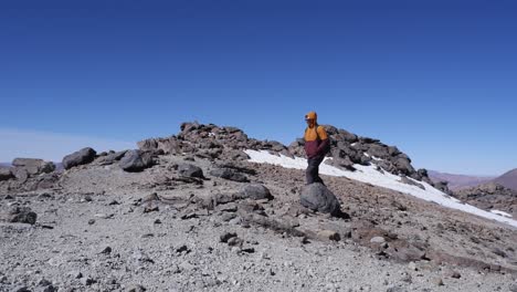 Man-walks-on-mountain-summit-trail-past-snow-patch-against-blue-sky