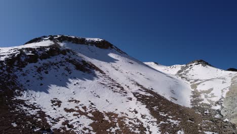 Aerial-rises-over-snowy-rock-slope-of-mountain-peak-against-blue-sky