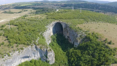 Retreating-drone-shot-of-the-wide-opening-of-Prohodna-Cave,-the-picturesque-view-of-the-mountains-and-woodlands-in-the-background,-located-in-Karlukovo,-in-Bulgaria