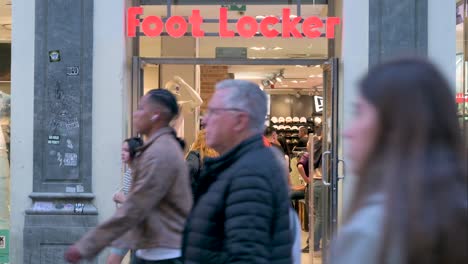 Shoppers-arrive-at-the-American-multinational-sportswear-and-footwear-retailer,-Foot-Locker,-store-as-pedestrians-walk-past-the-frame-at-a-busy-retail-street