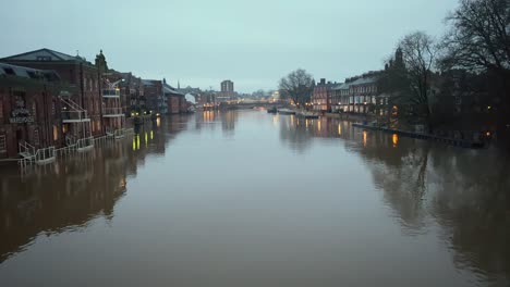 The-tranquil-yet-swollen-York-River-Ouse-reflects-the-evening-sky-as-the-water-level-rises,-inundating-the-riverside-walkways-up-to-the-building's-lower-levels