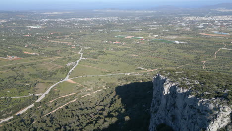 Drone-footage-near-Athens,-Greece,-showcasing-a-cliffside-flight-with-greenery-below-and-a-distant-Greek-town