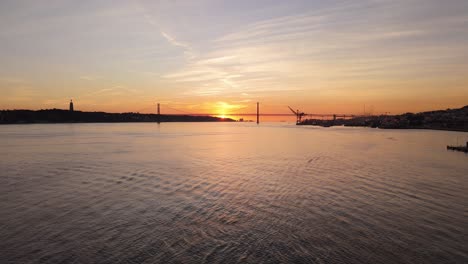 Niedrige-Panorama-Luftfahrt-Mit-Dolly-Entlang-Eines-Faszinierenden-Flussmusters-Mit-Hängebrückensilhouette-Bei-Sonnenuntergang