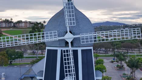 Close-up-on-center-of-windmill-in-Carlsbad,-pulling-back-to-reveal-cloudy-morning-sky-and-green-flower-fields-by-drone